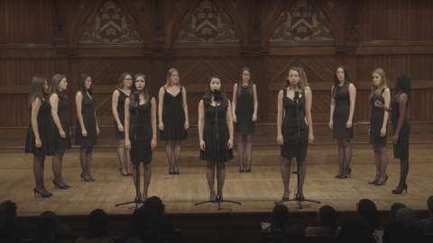 group of women singers in black dresses standing on stage, with three women standing in front of them behind mics.