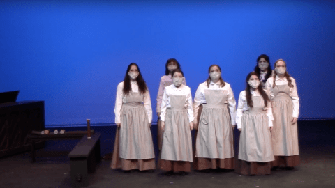 two rows of women actors wearing aprons and working clothes standing and singing.