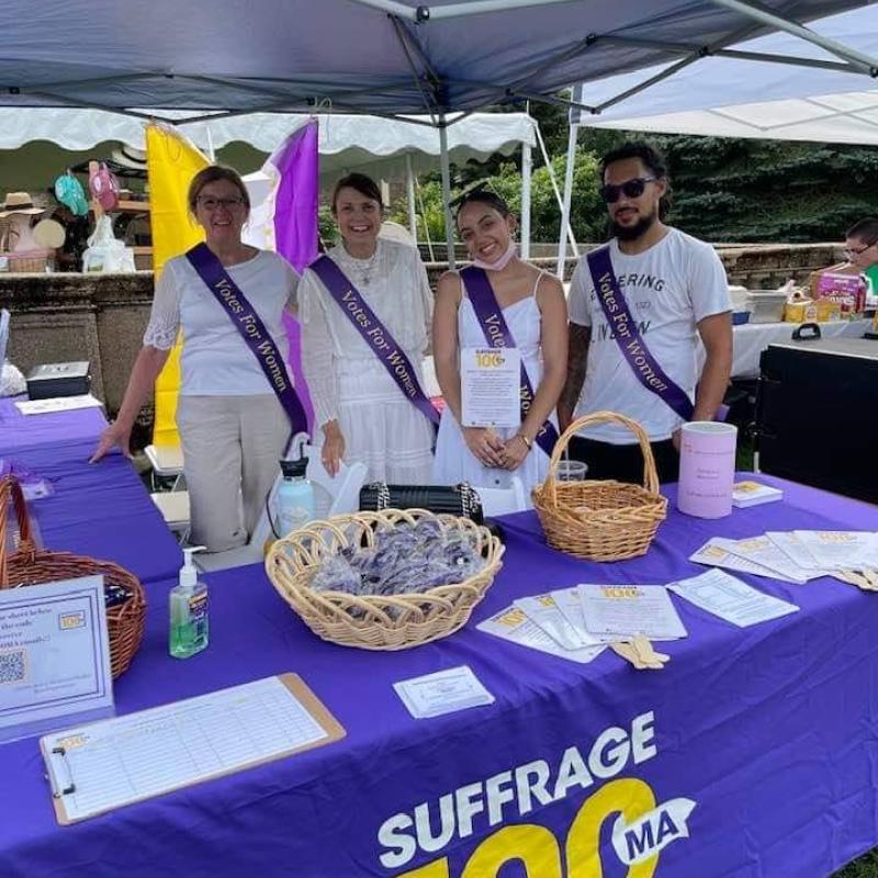 Four women stand outside at purple table.