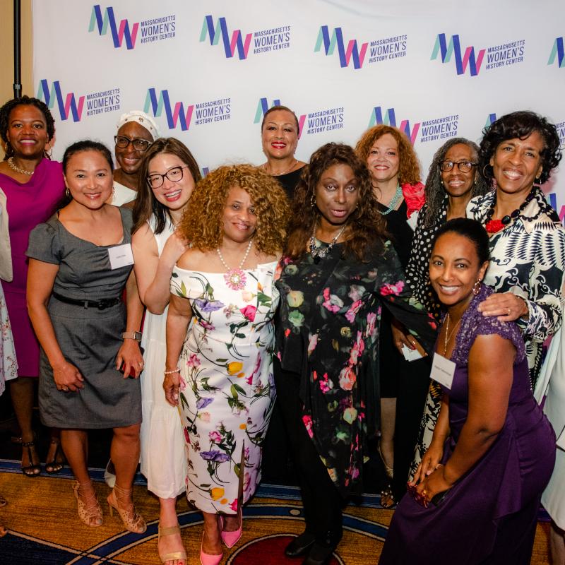 Large group of women stand in front of Massachusetts Women's History Center's step and repeat.