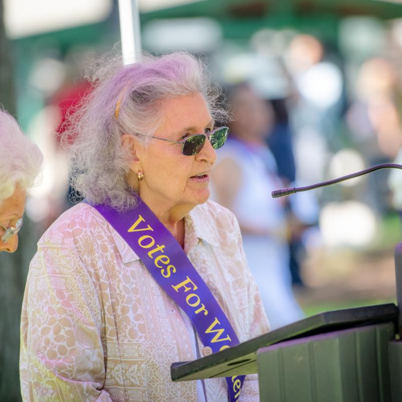 Woman wearing purple sash stands outside speaking at lectern.