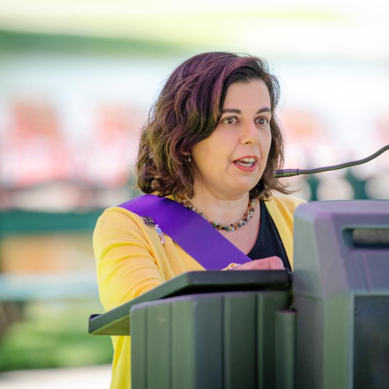 Woman wearing purple sash stands outside speaking at lectern.