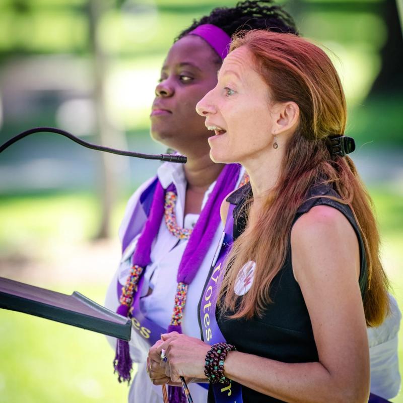 Two women wearing purple sash stand outside speaking at lectern.