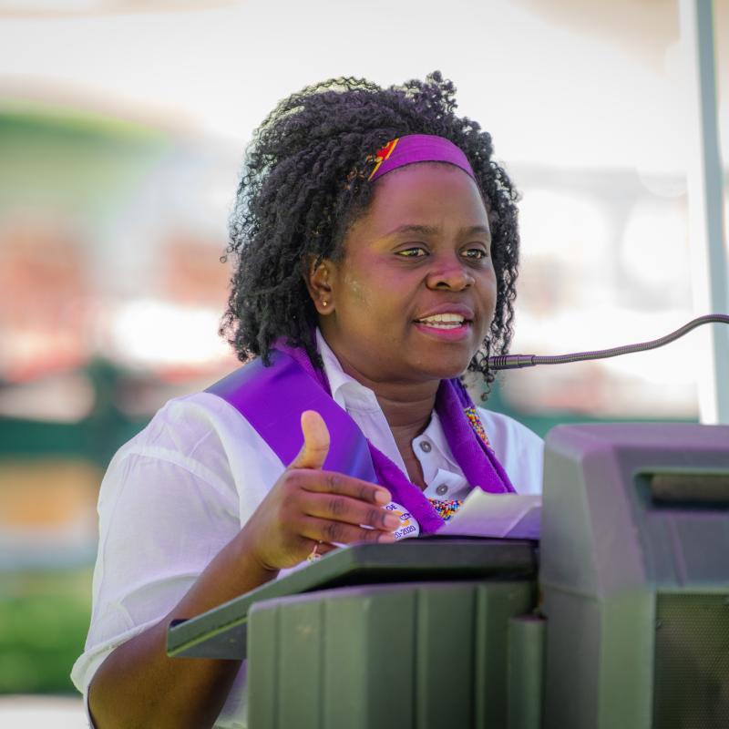 Woman wearing purple sash stands outside speaking at lectern.