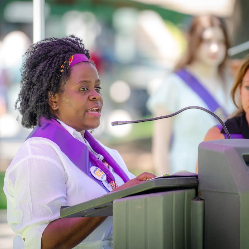 Woman wearing purple sash stands outside speaking at lectern.