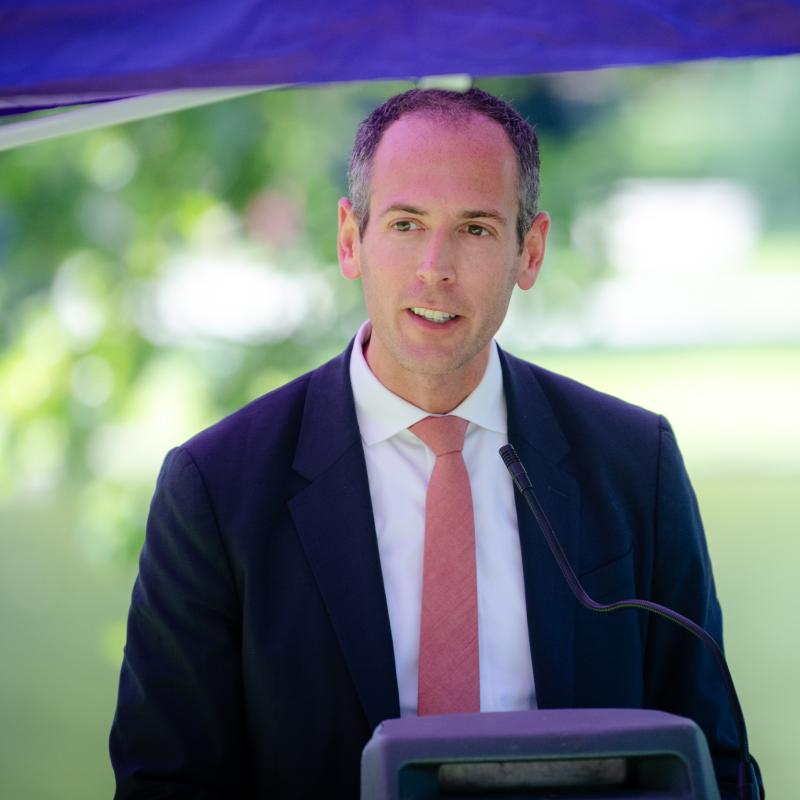 Man wearing purple sash stands outside speaking at lectern.