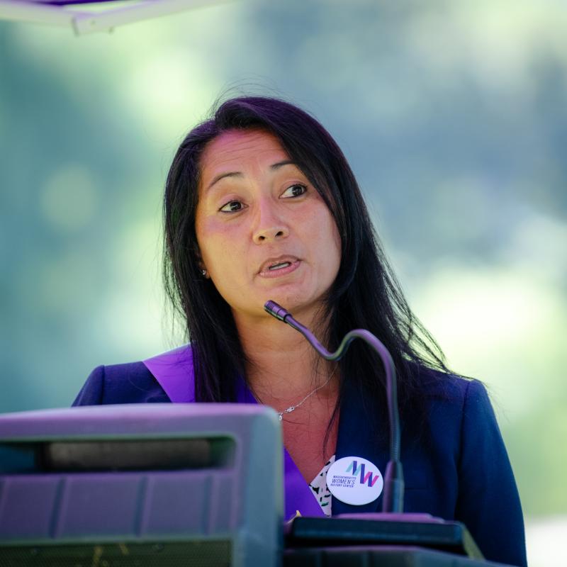 Woman wearing purple sash stands outside speaking at lectern.