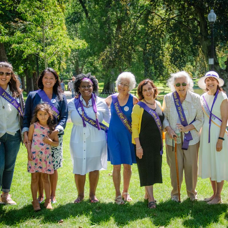 Group of women wearing purple sashes stand outside smiling.
