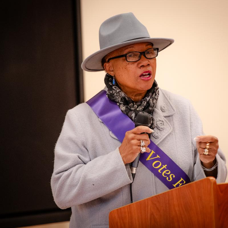 Woman wearing purple sash speaking at podium.