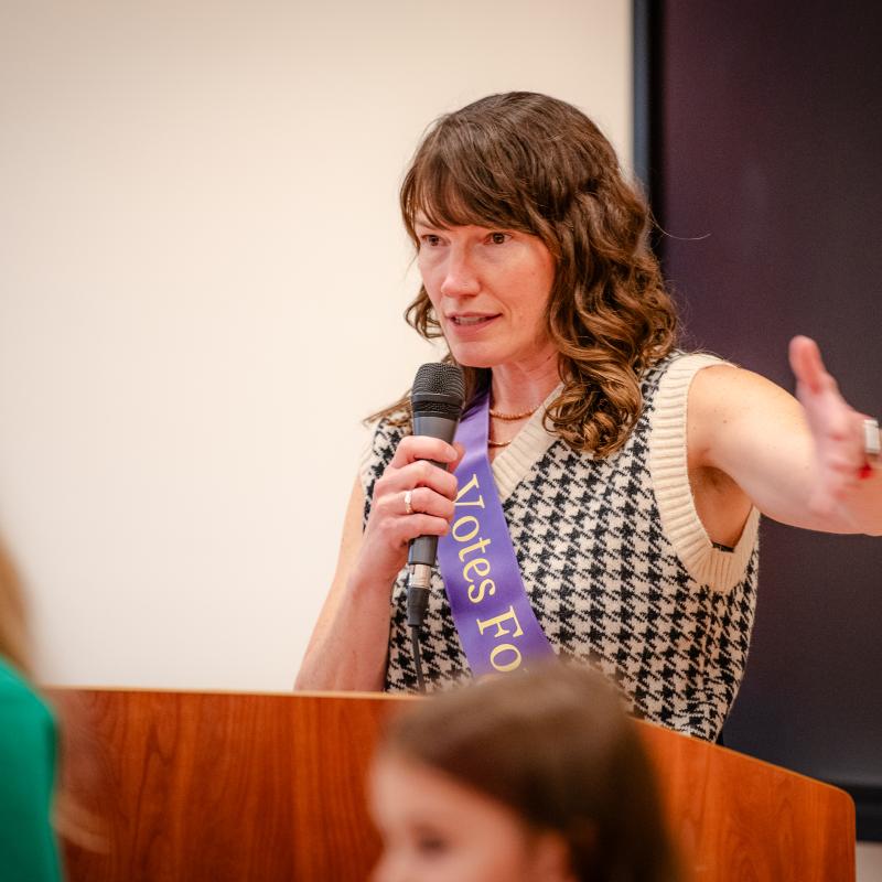 Woman wearing purple sash sits outside smiling.