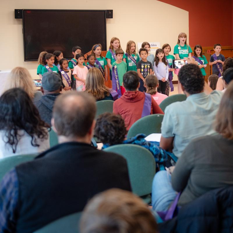 Group of children stand singing in front of crowd.
