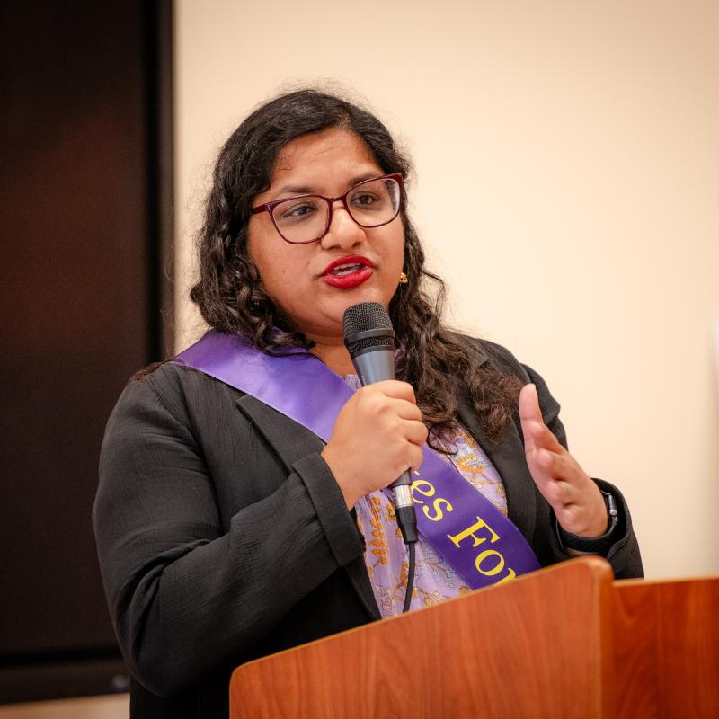 Woman wearing purple sash stands at podium holding microphone.