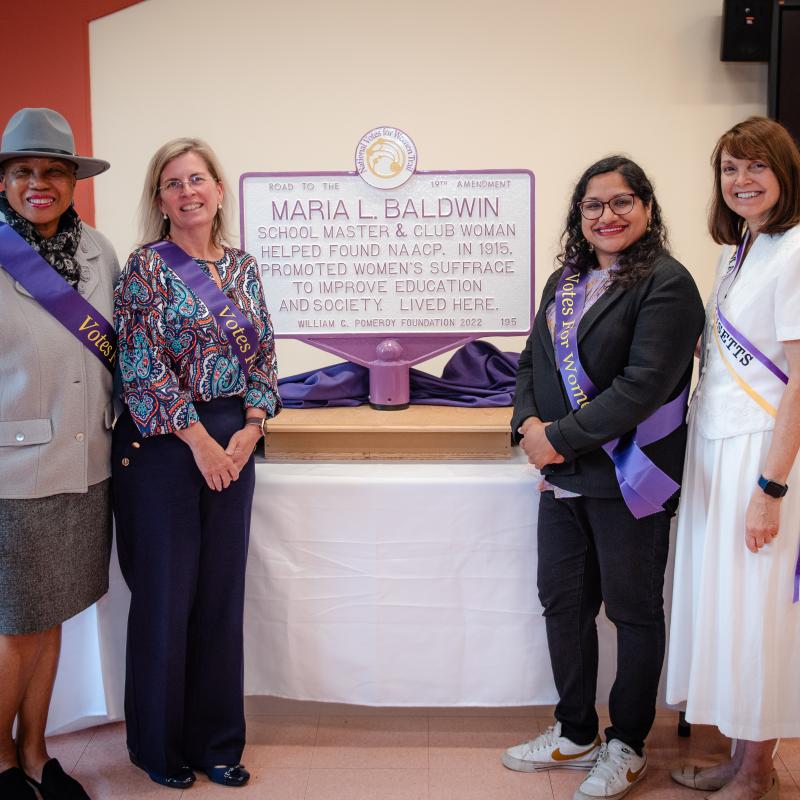 Four women wearing purple sashes stand inside in front of white and purple sign.