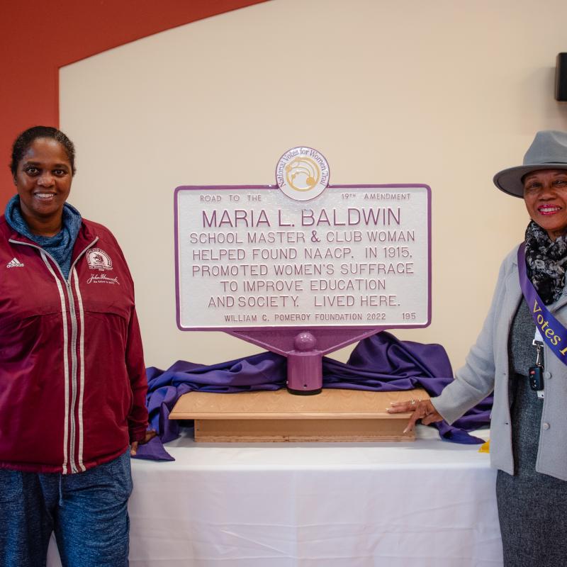 Two people wearing purple sashes stand smiling in front of purple and white sign.