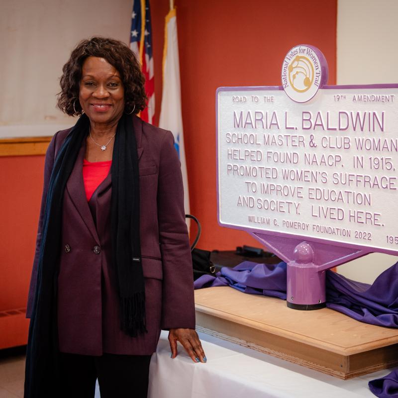 Woman stands next to purple and white sign.