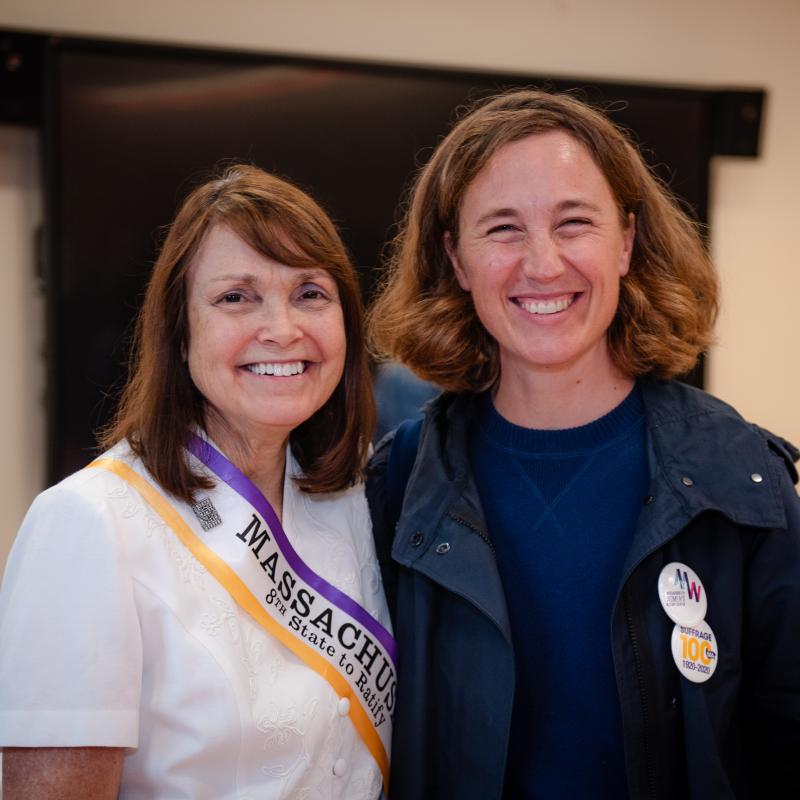 Two women stand smiling indoors.