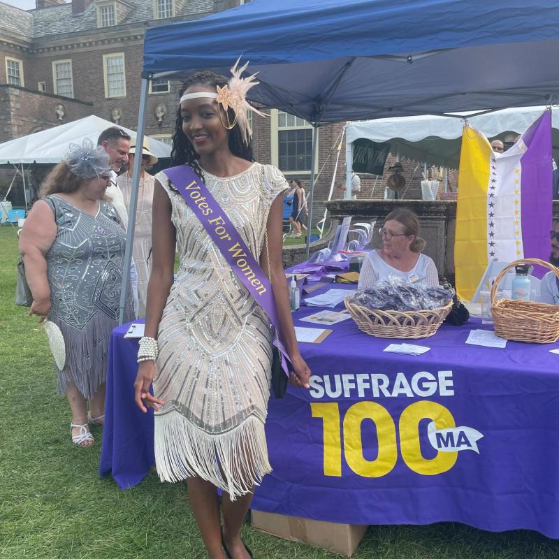 Woman stands outside wearing sash.