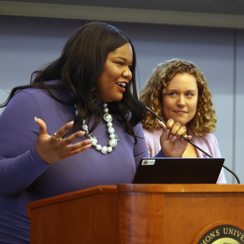 Woman speaks at podium while second woman watches.