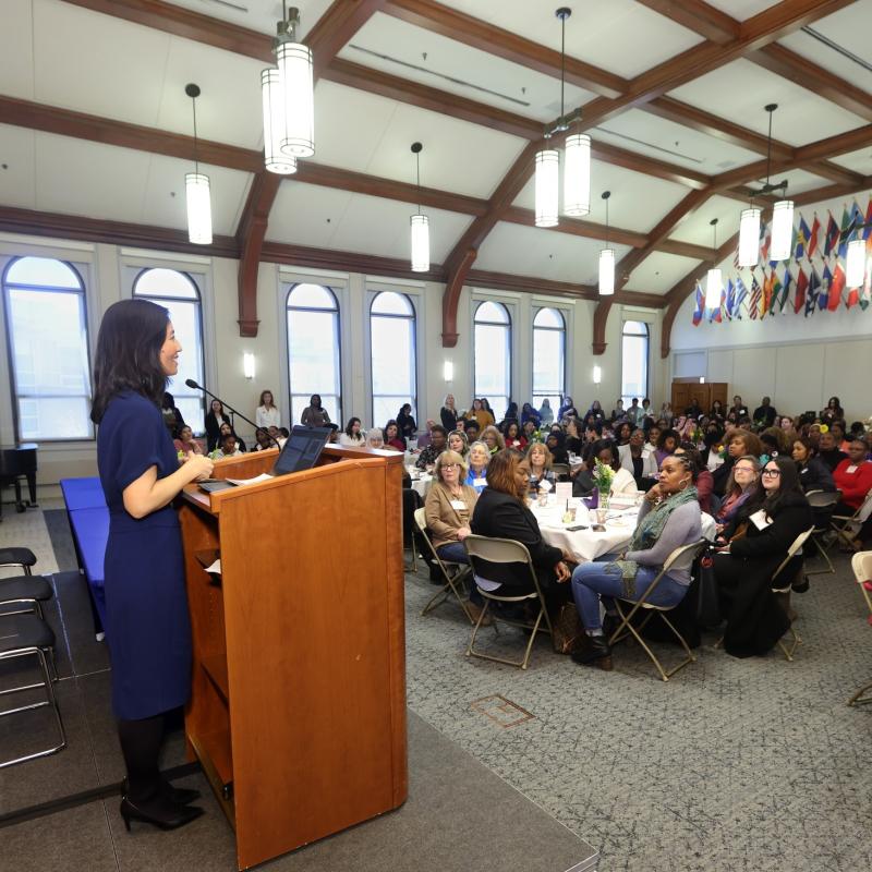 Woman speaks at podium in front of large seated crowd.