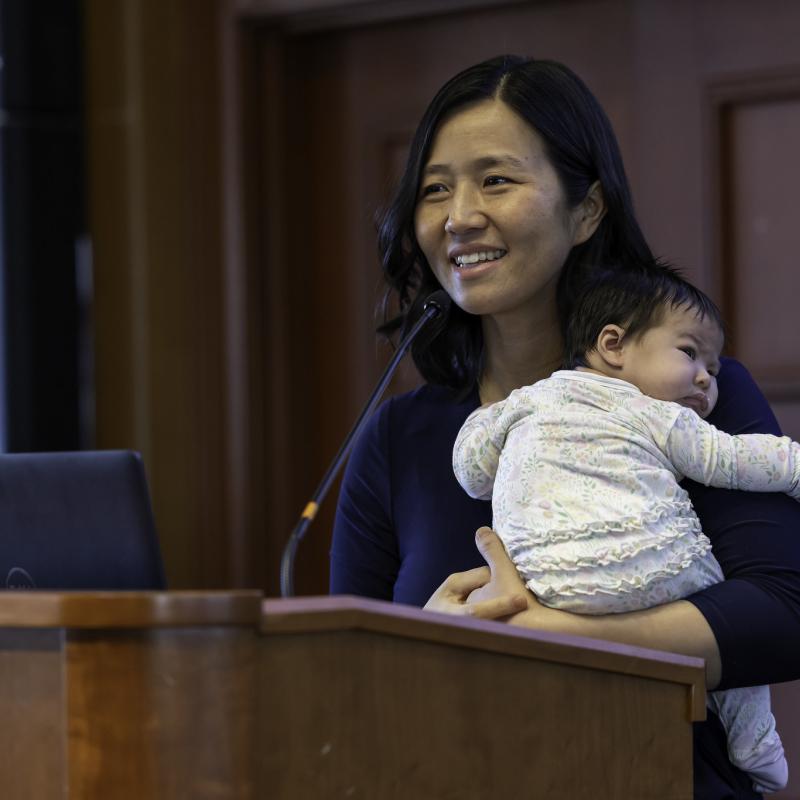 Mayor Michelle Wu holding a baby and addressing a group from a podium
