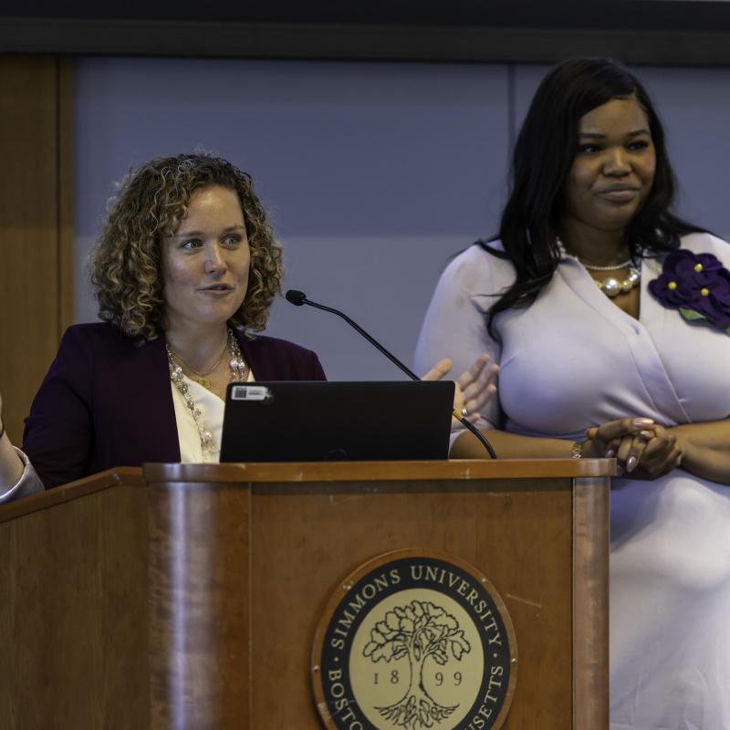 A woman speaking from a podium and anothe woman standing behind her smiling