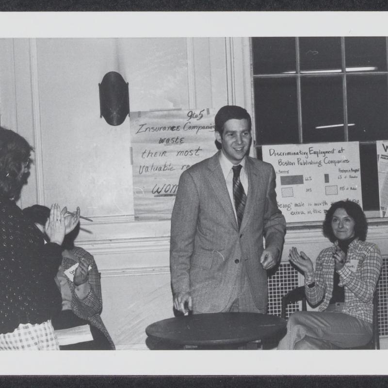 Man wearing a suit and tie stands in front of a 9to5 poster. A few women are sitting or standing around him and clapping.