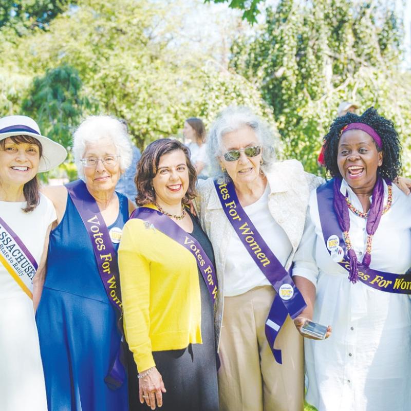 Six women wearing purple sashes smile outdoors.