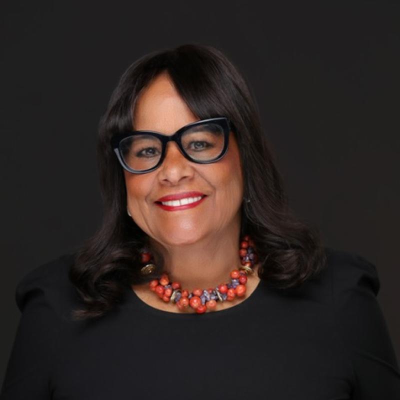 headshot of a woman smiling and wearing a black jacket, glasses, and red necklace