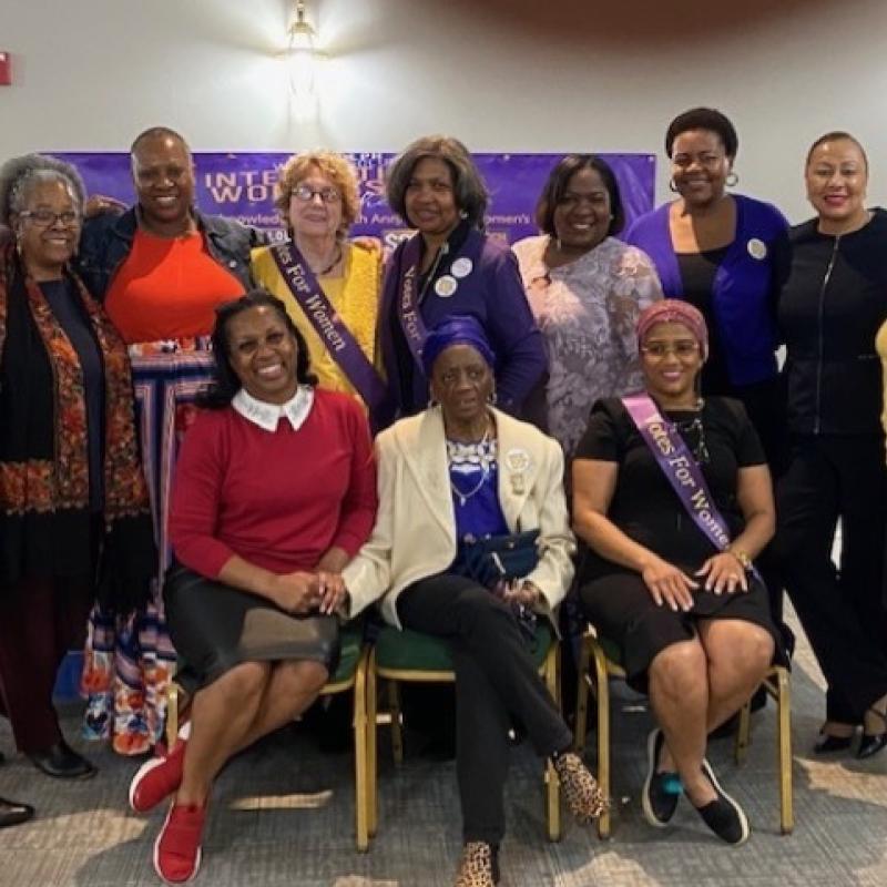 Group of women standing and sitting, wearing purple sashes.