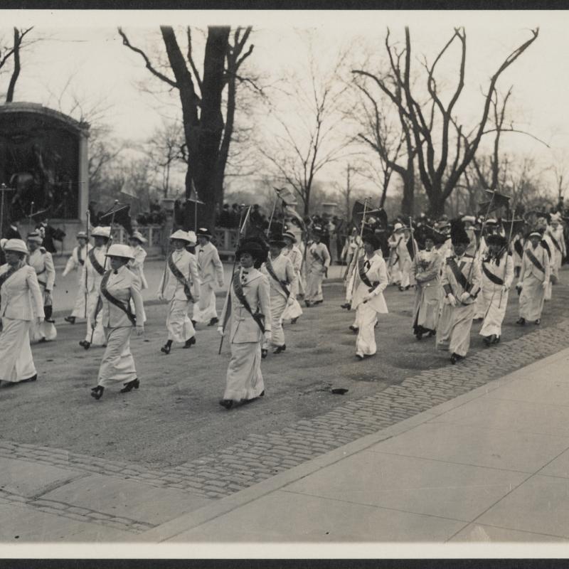 A regiment of women in uniform (white or pale clothes and a dark sash) carrying staffs and marching in formation in a suffrage parade. Crowds look on from both sidewalks.