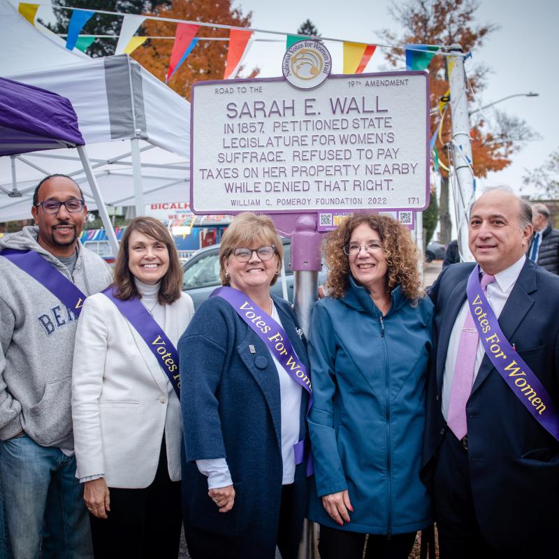 Four people stand outside in front of white and purple sign.