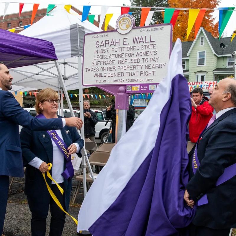 Group stands outside looking at white and purple sign.
