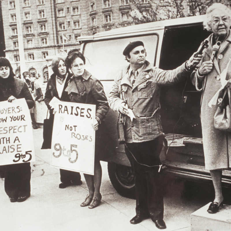 older woman wearing a coat stands on a box as a man holds a microphone up to her. Two other women stand with signs nearby.