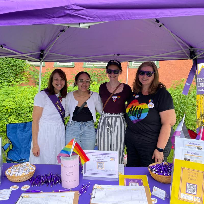 Four women stand at purple table.