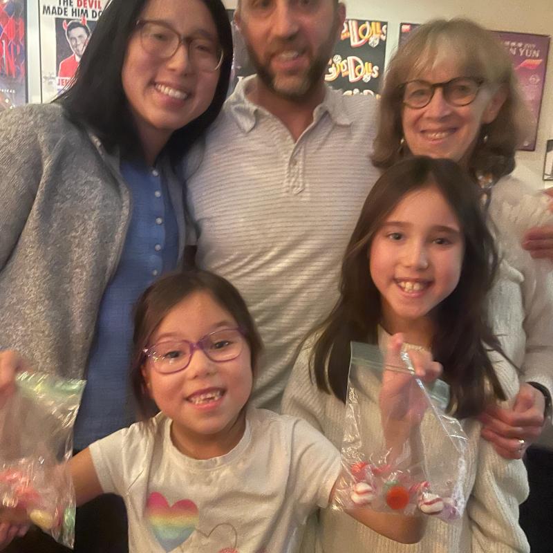 A group of five people, including two adults and three children, are smiling at the camera. The two children in front are holding small bags with striped candies. Posters are visible in the background.