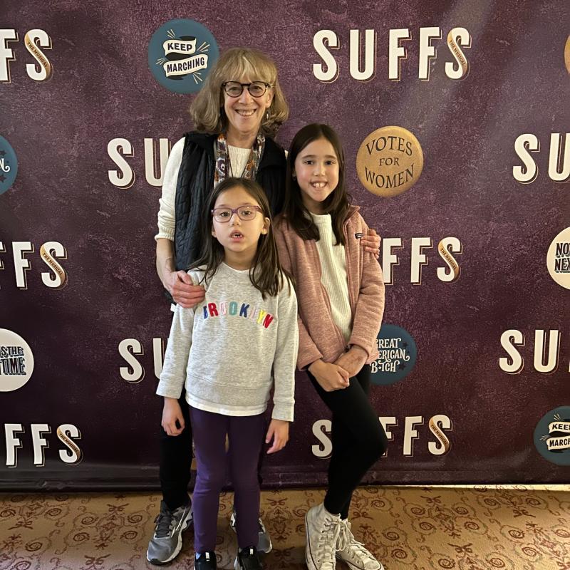 A woman and two children in front of a "Votes for Women" themed backdrop.