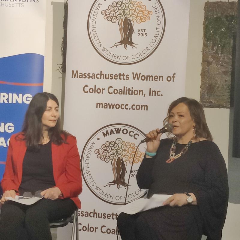 Two women sit in folding chairs, one is holding a microphone.
