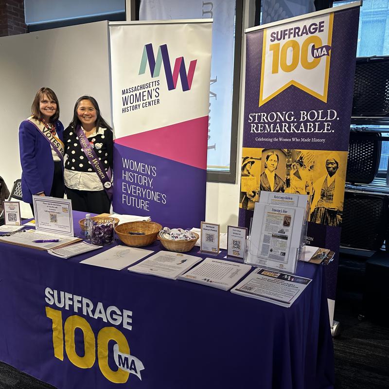Two women stand indoors at purple table.