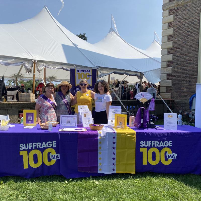Four women stand outside behind purple table.