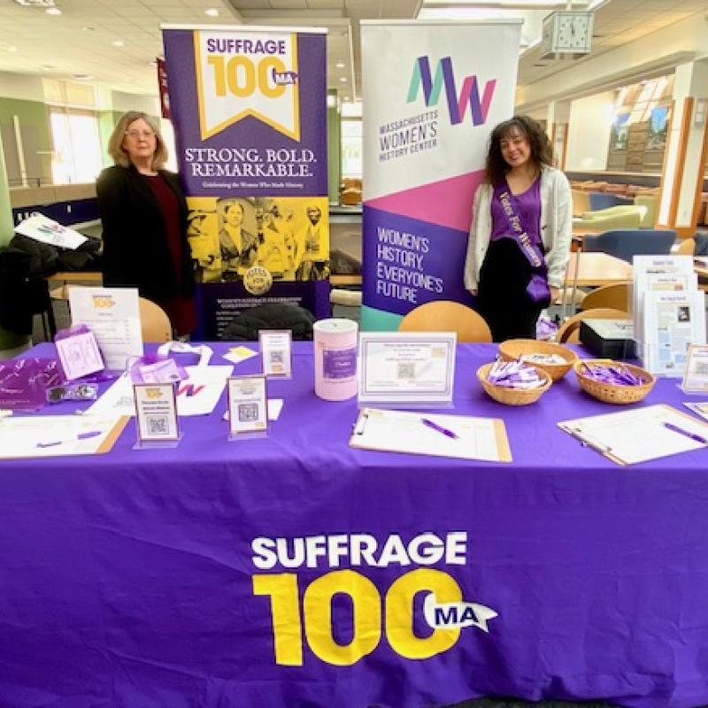 Two women stand at purple table.