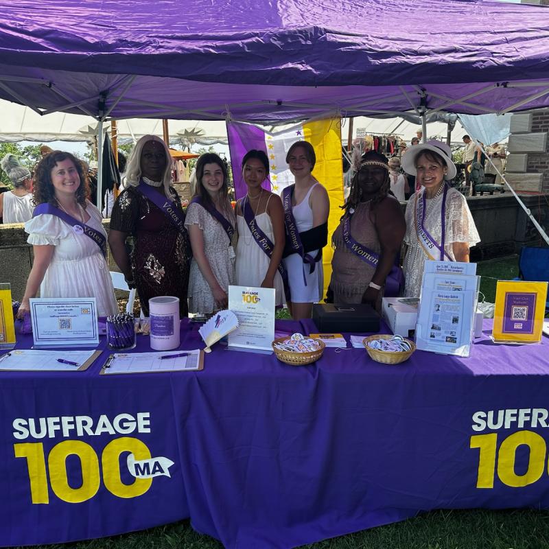 Seven women stand outside behind purple table.