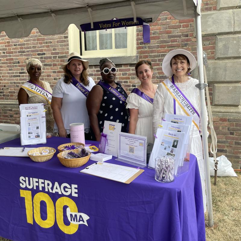Five women stand outside at purple table.