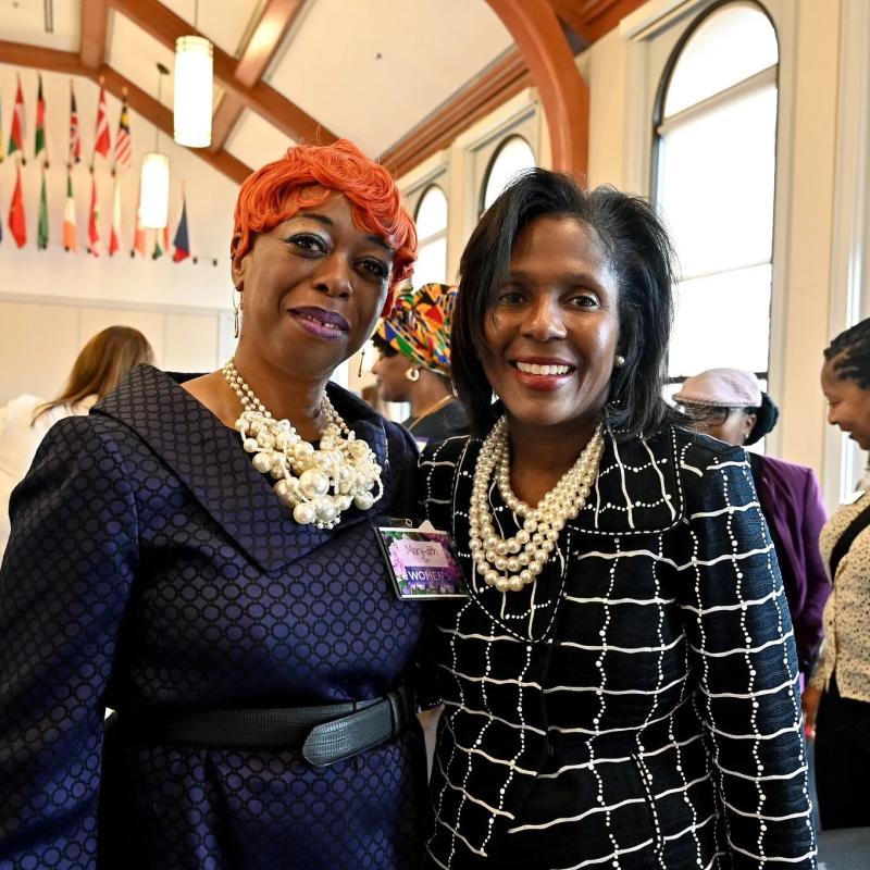 Two women stand indoors smiling.