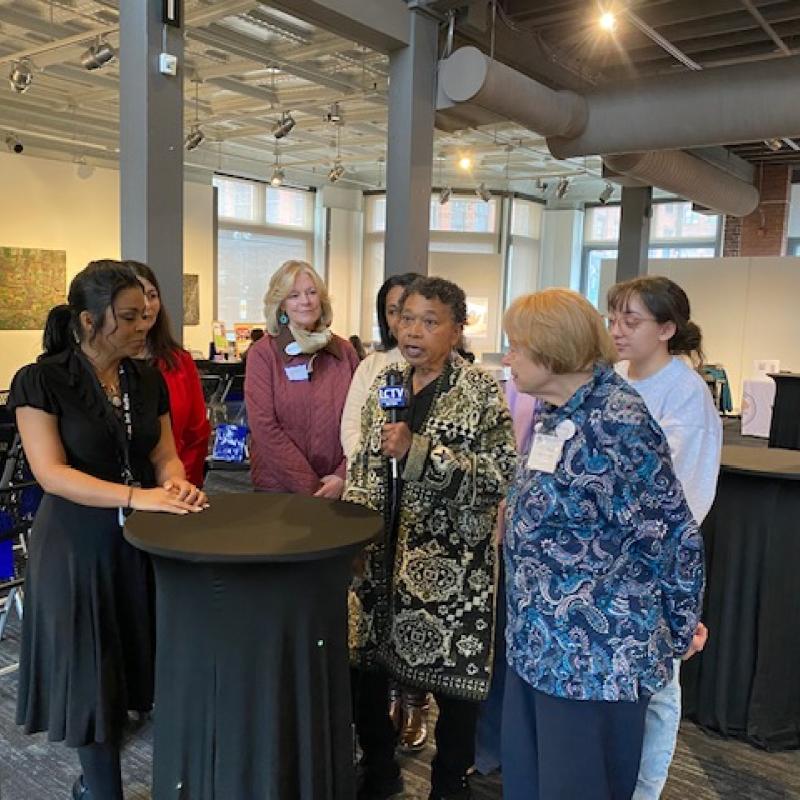 Five women stand at black table speaking to each other.
