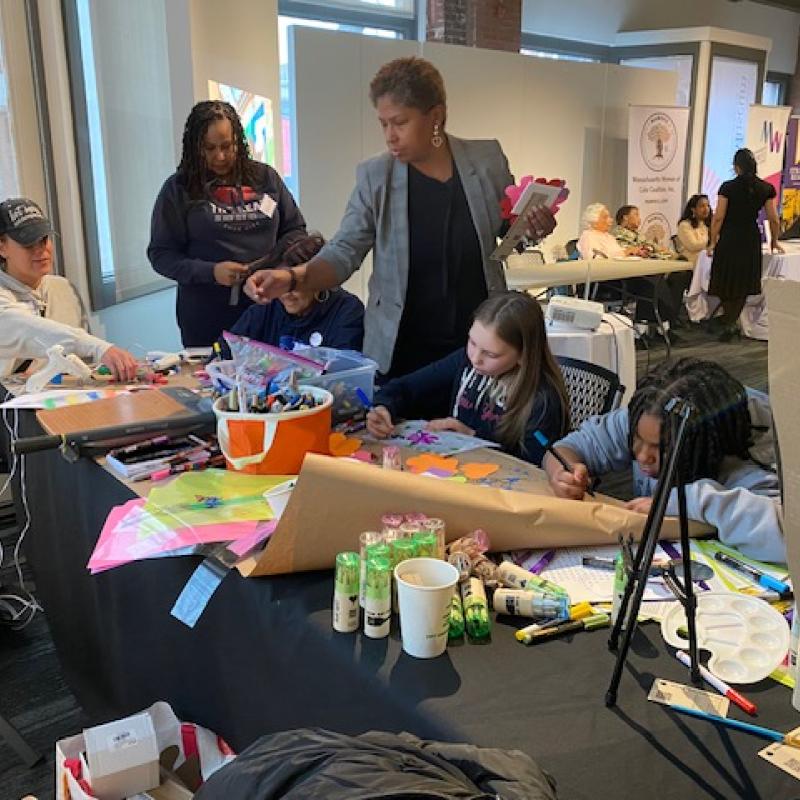 Three girls do crafts at table while two women look on.