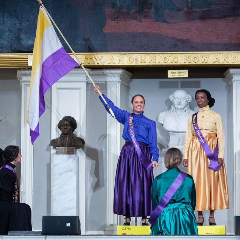 Two women stand on stage wearing purple sashes; one woman holds flag with stripes of gold, purple and white.
