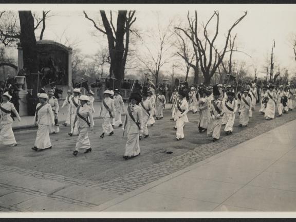 A regiment of women in uniform (white or pale clothes and a dark sash) carrying staffs and marching in formation in a suffrage parade. Crowds look on from both sidewalks.