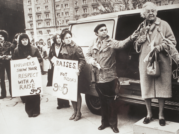older woman wearing a coat stands on a box as a man holds a microphone up to her. Two other women stand with signs nearby.