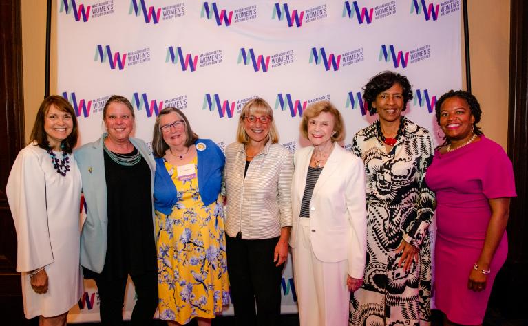 Seven women stand in front of Massachusetts Women's History Center's step and repeat.