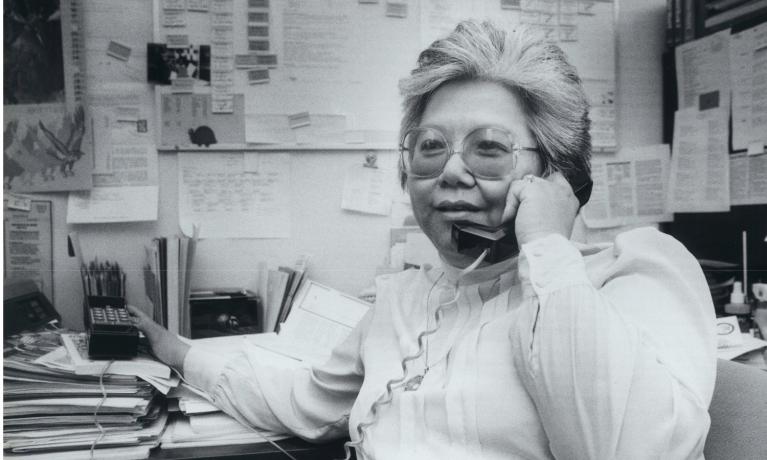 Black and white photo of woman sitting at desk, speaking on phone.